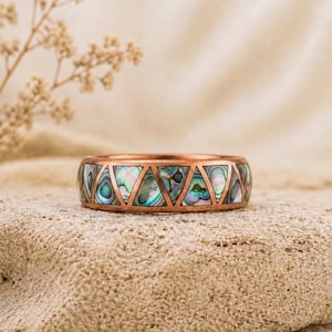 May include: A copper ring featuring a series of triangular inlays made of iridescent abalone shell. The metal band has a smooth finish and a geometric pattern that alternates between solid copper triangles and colorful shell segments.