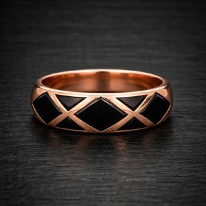 May include: A rose gold ring with a geometric pattern. The ring has a series of black diamond shapes set within a rose gold band. The ring is shown on a dark surface.