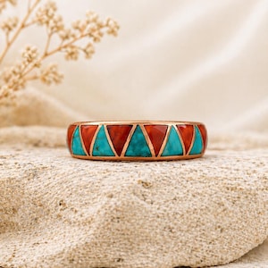 May include: A copper ring featuring a geometric pattern of turquoise and red triangular inlays. The ring is presented on a textured, light-toned surface. The ring's design is eye-catching and bold.