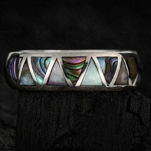 May include: A silver ring with a geometric design. The ring features triangular cutouts filled with iridescent abalone shell. The ring is displayed on a dark, textured surface, showcasing its intricate details and craftsmanship.