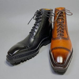 May include: Two leather boots, one black and one tan, are displayed side-by-side. The black boot has a glossy finish and textured leather. The tan boot has a lighter, polished appearance. Both boots have laces and a square toe.
