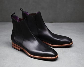 Pode incluir: Um par de botas Chelsea de couro preto. Estas botas apresentam um design elegante com painéis laterais elásticos e uma presilha. As solas são castanhas com uma borda clara, e o interior é forrado com tecido roxo.
