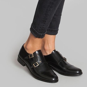 May include: Black leather double-buckle shoes with gold-colored hardware. The shoes have a polished finish and a low heel. The buckles are square and secure the straps across the top of the foot. The shoes are worn with black denim jeans.