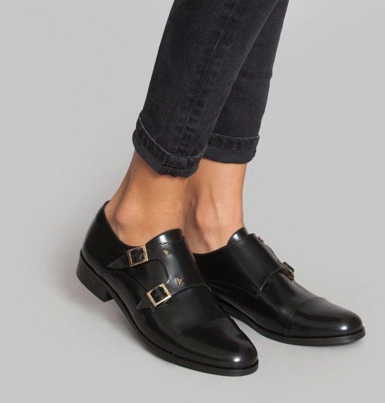 May include: Black leather double-buckle shoes with gold-colored hardware. The shoes have a polished finish and a low heel. The buckles are square and secure the straps across the top of the foot. The shoes are worn with black denim jeans.