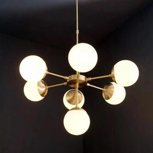 Op de afbeelding: Een moderne kroonluchter met een messing afwerking en meerdere bolvormige, matglazen kappen. De lamp heeft een centrale naaf met uitstralende armen, elk met een bol. Het ontwerp is minimalistisch en elegant.