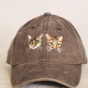 May include: A brown baseball cap featuring embroidered cat faces. The cap has a curved brim and a fabric strap closure. The cat faces are detailed with various colors, including brown, tan, and white.