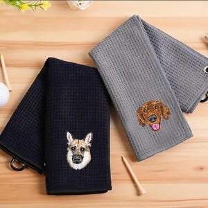 May include: Three golf towels with embroidered dog faces. One navy blue towel features a German Shepherd, while the other two are gray with a Golden Retriever. Each towel has a waffle weave texture and a metal ring for hanging.