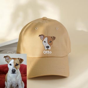 May include: A tan baseball cap featuring an embroidered image of a dog's face with brown and white markings. The dog's name, "Otto," is embroidered below the image in white letters. The cap has a curved brim and a button on top.