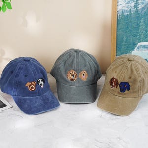May include: Three baseball caps in blue, gray, and tan, each embroidered with dog faces. The blue cap has a brown and a black and white dog, the gray cap has two golden retrievers, and the tan cap has a brown and a blue dog.