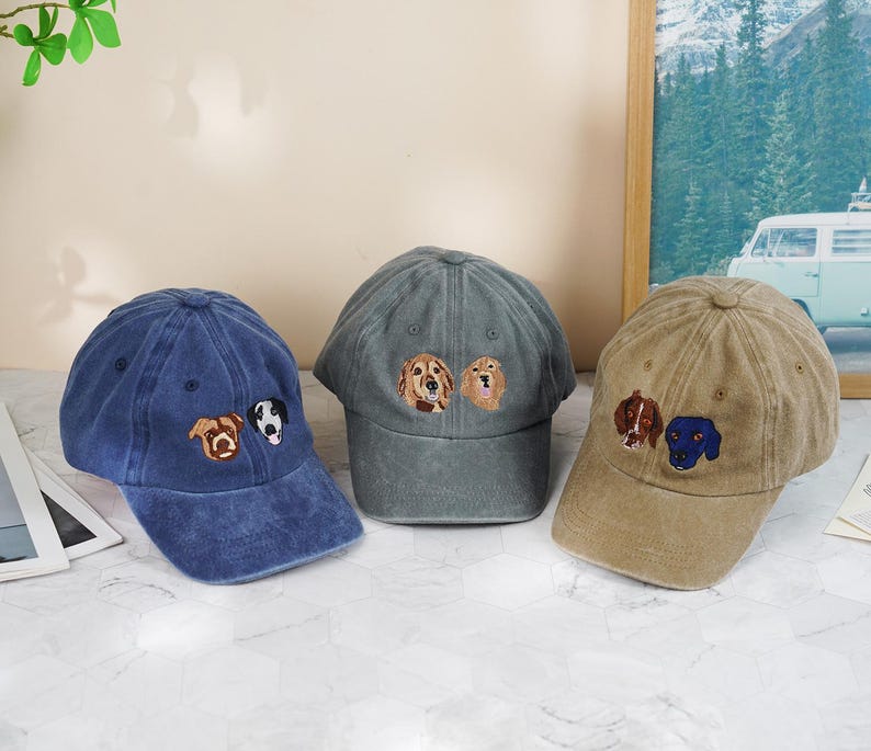 May include: Three baseball caps in blue, gray, and tan, each embroidered with dog faces. The blue cap has a brown and a black and white dog, the gray cap has two golden retrievers, and the tan cap has a brown and a blue dog.