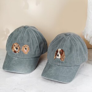 May include: Two gray baseball caps with embroidered dog faces. One cap features two golden retriever faces, and the other has a spaniel face. The caps have a curved brim and a classic six-panel design.