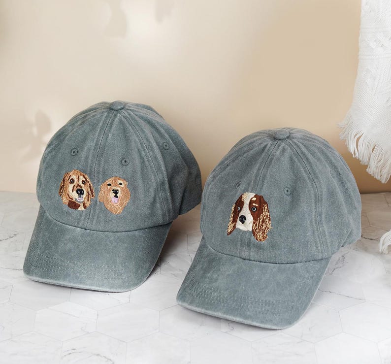 May include: Two gray baseball caps with embroidered dog faces. One cap features two golden retriever faces, and the other has a spaniel face. The caps have a curved brim and a classic six-panel design.