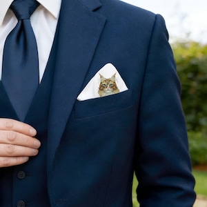 May include: A navy blue suit with a matching tie and a white pocket square featuring a detailed illustration of a cat. The cat has brown and tan fur with blue eyes. The suit has a three-button closure.