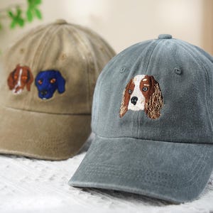 May include: Two baseball caps with embroidered dog faces. One cap is tan with two dog faces, and the other is a muted teal with a single dog face. The dog faces are brown, white, and blue.