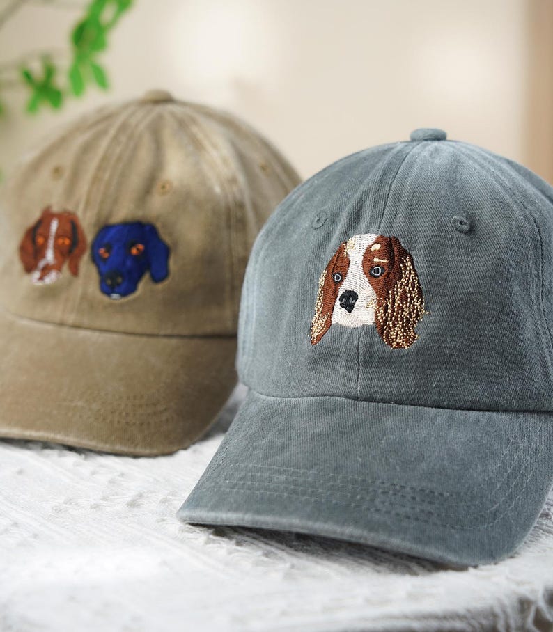 May include: Two baseball caps with embroidered dog faces. One cap is tan with two dog faces, and the other is a muted teal with a single dog face. The dog faces are brown, white, and blue.