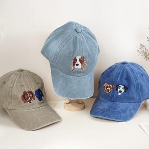 May include: Three baseball caps in shades of blue and tan, each embroidered with a dog's face. The caps are displayed on a light-colored surface, showcasing the detailed canine designs.