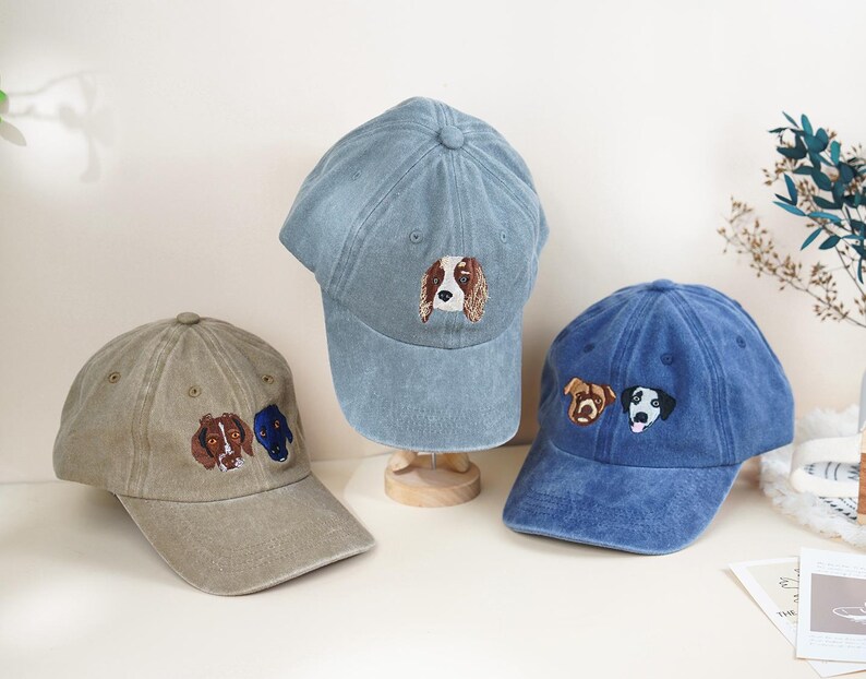 May include: Three baseball caps in shades of blue and tan, each embroidered with a dog's face. The caps are displayed on a light-colored surface, showcasing the detailed canine designs.