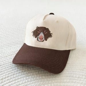 Puede incluir: Gorra de béisbol con visera marrón y corona color crema. La gorra presenta una imagen bordada de la cara de un perro en marrón y rosa. La gorra tiene una visera curva y un botón en la parte superior.