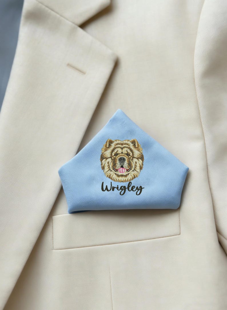May include: A pale blue pocket square featuring an embroidered Chow Chow dog's face and the name "Wrigley" in black script. The pocket square is folded and placed in the breast pocket of a cream-coloured blazer.