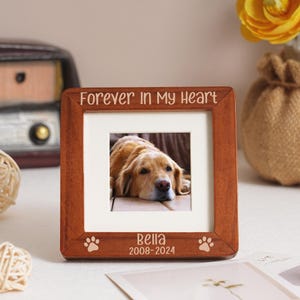 May include: A square, wooden photo frame with the inscription "Forever In My Heart" at the top. The frame contains a photograph of a golden retriever. The dog's name, "Bella", and the years "2008-2024" are printed at the bottom of the frame.