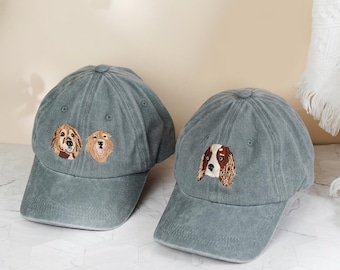 May include: Two gray baseball caps with embroidered dog faces. One cap features two golden retriever faces, and the other has a spaniel face. The caps have a curved brim and a classic six-panel design.