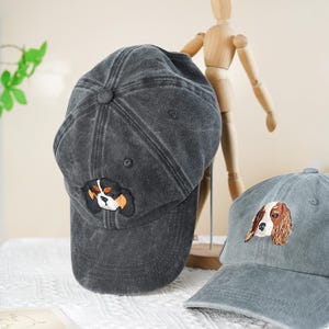May include: Two gray baseball caps with embroidered dog faces. One cap is a darker gray, the other is a lighter gray. The caps have a curved brim and a fabric strap closure. The dog embroidery features black, white, and brown colors.