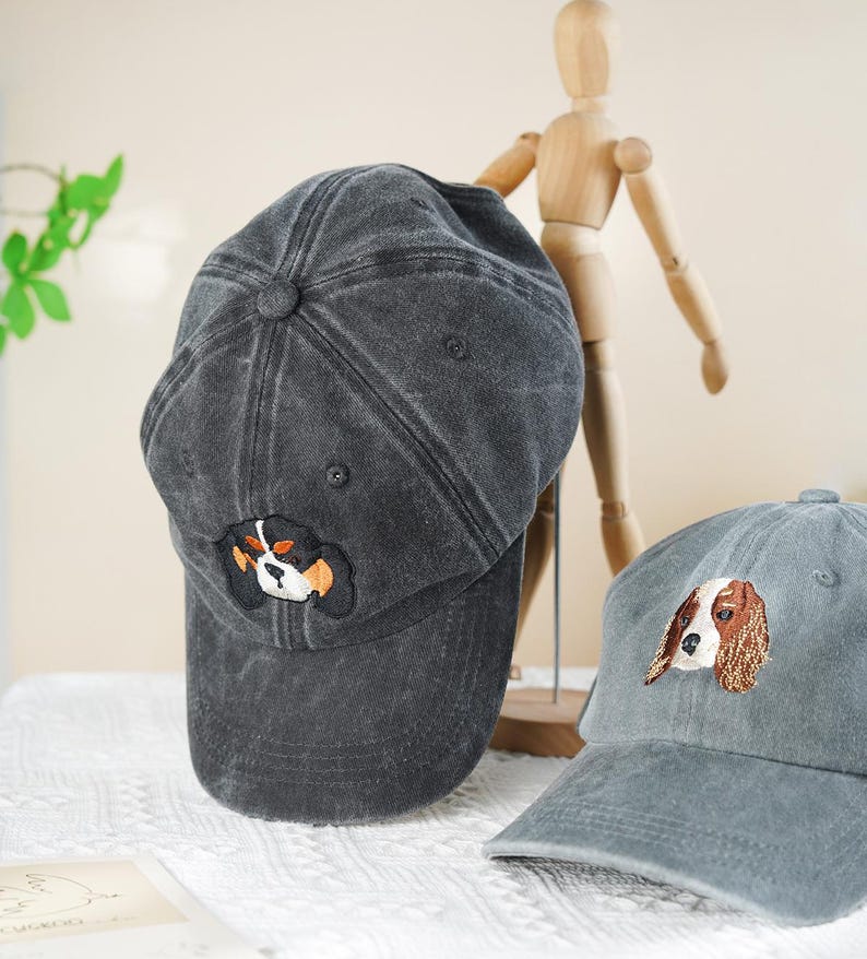May include: Two gray baseball caps with embroidered dog faces. One cap is a darker gray, the other is a lighter gray. The caps have a curved brim and a fabric strap closure. The dog embroidery features black, white, and brown colors.