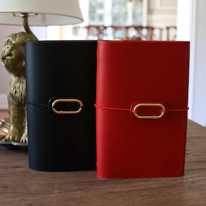 May include: Two leather journals, one black and one red, stand side by side. Each journal features a gold-colored clasp and a matching elastic band closure. The journals are displayed on a wooden surface.
