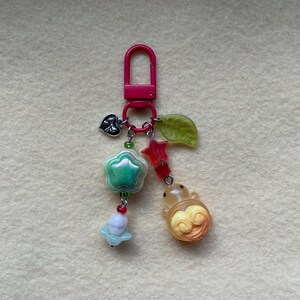 May include: A pink keychain with various charms. Charms include a green flower, a red star, a yellow swirl, a heart, and a leaf. The keychain is made of metal and plastic.