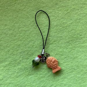 May include: A small charm with a black cord, featuring a peach-colored fish, a heart with a bow, a white bead, a green bead, and a red heart. The charm is attached to a silver-colored metal loop.