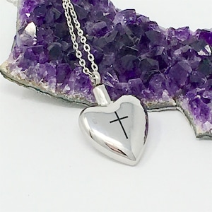 May include: A silver heart-shaped pendant necklace with a cross engraved on the front. The necklace is displayed on a purple amethyst crystal.