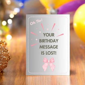 May include: A silver birthday card with the text "Oh No! Your Birthday Message Is Lost!" in black and pink. The card features a pink bow and decorative lines. The background is blurred with gold and yellow.