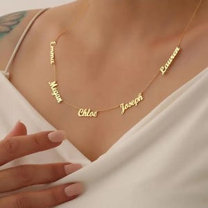 Op de afbeelding: Een goudkleurige ketting met meerdere naamhangers. De namen "Emma", "Megan", "Chloe", "Joseph" en "Lauren" worden weergegeven. De ketting wordt om de nek gedragen.