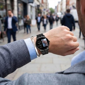 May include: A black smartwatch with a matching metal band displays a photo and text. The text reads "Our Love Between Father & Daughter Lives Forever." A silver bracelet with Roman numerals is also visible.