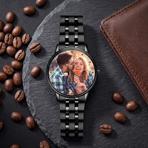 K&ouml;nnte beinhalten: Schwarze Armbanduhr mit Metallarmband. Das Zifferblatt zeigt ein Farbfoto eines Paares. Die Uhr liegt auf einer dunkelgrauen Oberfl&auml;che, auf der Kaffeebohnen verstreut sind. Im Hintergrund befindet sich eine braune Lederbrieftasche.