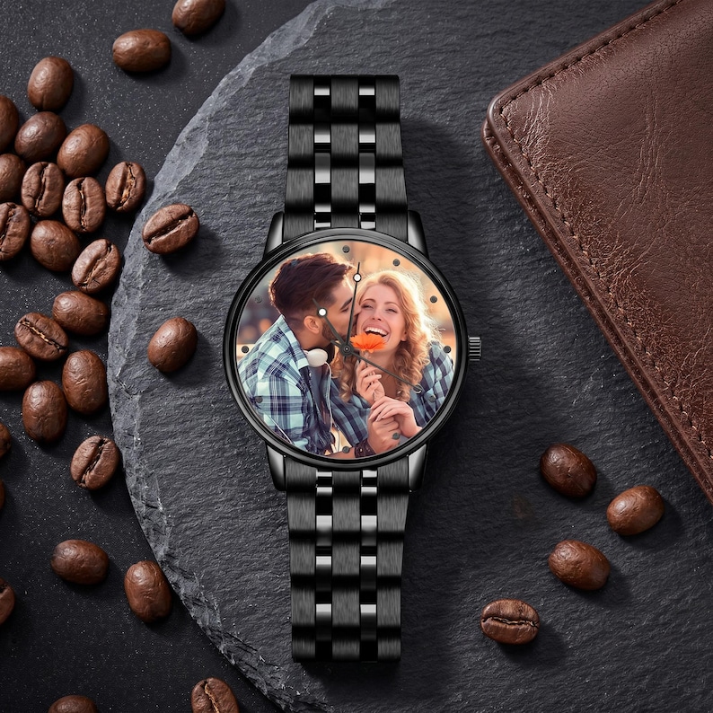 K&ouml;nnte beinhalten: Schwarze Armbanduhr mit Metallarmband. Das Zifferblatt zeigt ein Farbfoto eines Paares. Die Uhr liegt auf einer dunkelgrauen Oberfl&auml;che, auf der Kaffeebohnen verstreut sind. Im Hintergrund befindet sich eine braune Lederbrieftasche.