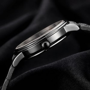 May include: A side view of a silver wristwatch with a black face and a silver band. The watch has a round face with a black bezel and a silver crown. The band is made of small, interconnected links. The watch is set against a dark background.
