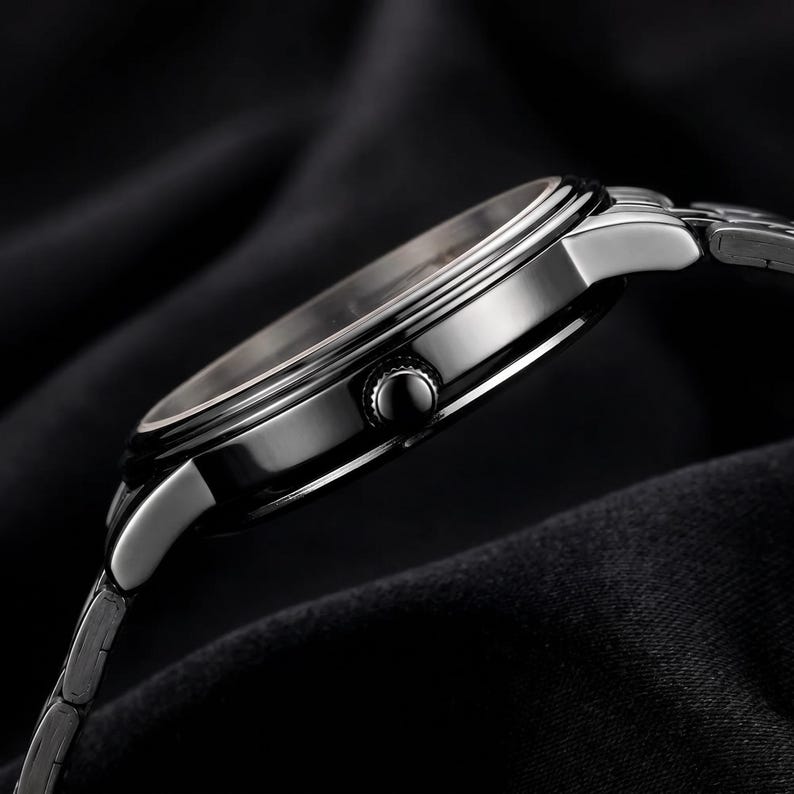 May include: A side view of a silver wristwatch with a black face and a silver band. The watch has a round face with a black bezel and a silver crown. The band is made of small, interconnected links. The watch is set against a dark background.