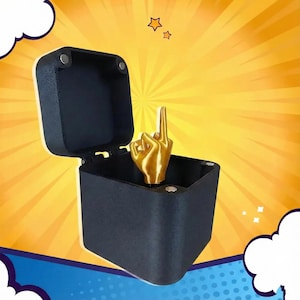 Puede incluir: Una escultura de mano dorada con el dedo medio levantado, expuesta dentro de una caja negra abierta. La caja tiene una tapa con bisagras y cierres magnéticos. El fondo es amarillo vibrante con líneas radiales y estrellas decorativas.