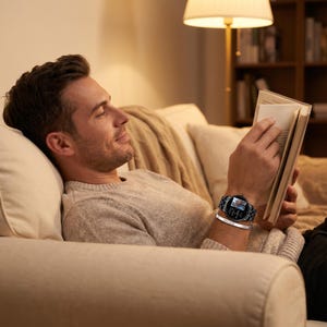 May include: A black smartwatch with a silver band and a silver bracelet. The watch has a round face with a digital display. The person is wearing a gray sweater and is reading a book while relaxing on a beige couch.