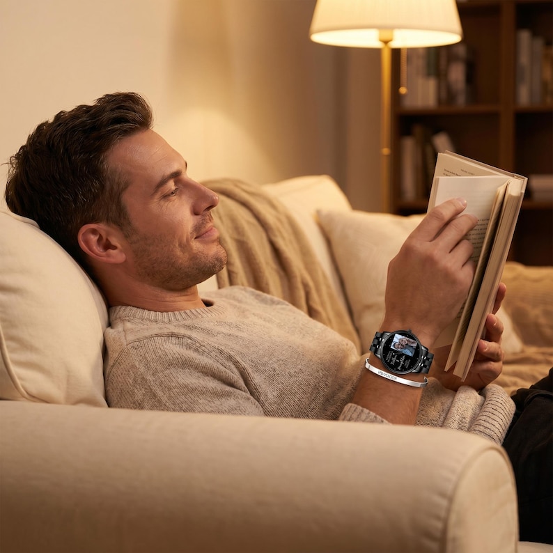 May include: A black smartwatch with a silver band and a silver bracelet. The watch has a round face with a digital display. The person is wearing a gray sweater and is reading a book while relaxing on a beige couch.