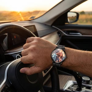 K&ouml;nnte beinhalten: Eine schwarze Armbanduhr mit Metallarmband wird am Handgelenk einer Person getragen. Das Zifferblatt zeigt ein Farbfoto eines Paares. Die Person h&auml;lt ein Lenkrad in einem Auto, mit einem Sonnenuntergang im Hintergrund.