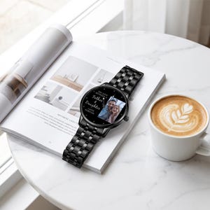May include: Black wristwatch with a metal band, resting on a magazine. The watch face displays a photo of a father and daughter, with the text "The Love Between a Father & Daughter Lasts Forever."
