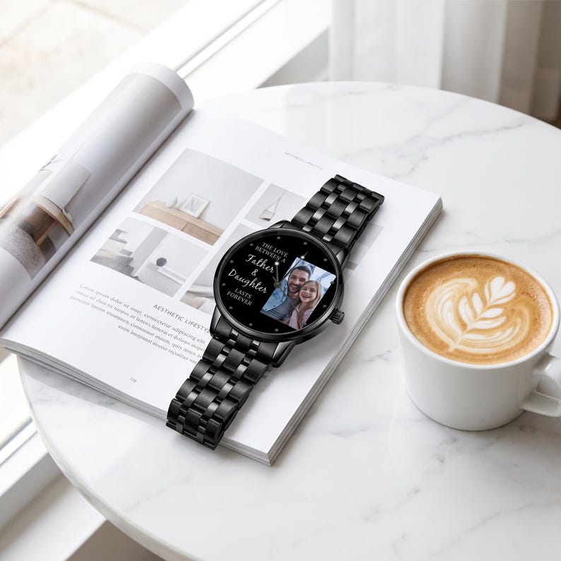 May include: Black wristwatch with a metal band, resting on a magazine. The watch face displays a photo of a father and daughter, with the text "The Love Between a Father & Daughter Lasts Forever."
