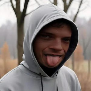May include: A light gray hooded sweatshirt with a black interior lining. The person is sticking their tongue out. The background is blurred with trees and a foggy atmosphere.