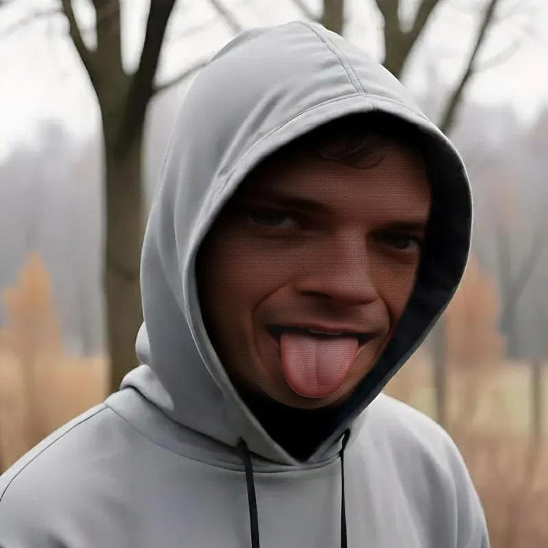 May include: A light gray hooded sweatshirt with a black interior lining. The person is sticking their tongue out. The background is blurred with trees and a foggy atmosphere.