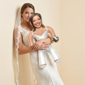Può includere: Una sposa in un abito da sposa di pizzo bianco abbraccia una bambola gonfiabile sorridente, anch'essa vestita con un abito da sposa bianco. La bambola tiene in mano un piccolo bouquet. La sposa indossa un velo e un abito di pizzo abbinato.