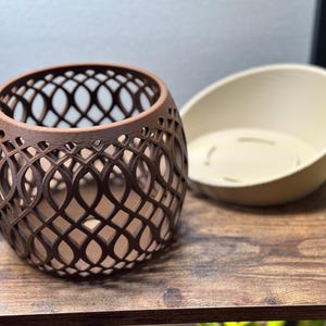 May include: A brown, openwork, spherical planter with a decorative lattice design. Beside it is a cream-colored, oval-shaped planter insert. The items are on a wooden surface.