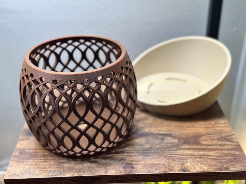 May include: A brown, openwork, spherical planter with a decorative lattice design. Beside it is a cream-colored, oval-shaped planter insert. The items are on a wooden surface.