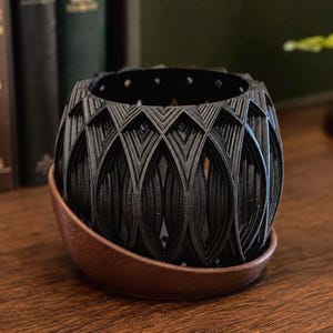 May include: A black, spherical, open-lattice planter with a repeating geometric pattern, resting inside a smooth, curved, copper-colored base. The planter features a hollow, cage-like structure with vertical ribs and a circular opening at the top.